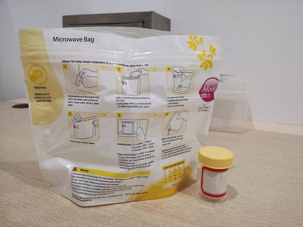 This clinical photograph displays a medical-grade disinfection system used for neonatal care accessories, specifically for the steam sterilization of breast milk collection equipment. The image features a specialized microwave steam sterilization bag alongside a sterile collection cup. The bag is a flexible, translucent white pouch with yellow instructional graphics and a 'Hold here' safety zone. It displays a six-step illustrated guide for cleaning, adding water (60ml), and microwave heating times based on wattage. A tracking grid at the bottom is included to monitor the bag's 20-use lifecycle. Next to the bag is a cylindrical, transparent collection cup with a yellow screw-top lid and a red-bordered label for patient identification. This visual represents standard protocols for infection control in a Neonatal Intensive Care Unit (NICU) or home care setting, demonstrating the process of ensuring the microbiological safety of colostrum and breast milk storage containers through rapid steam treatment.