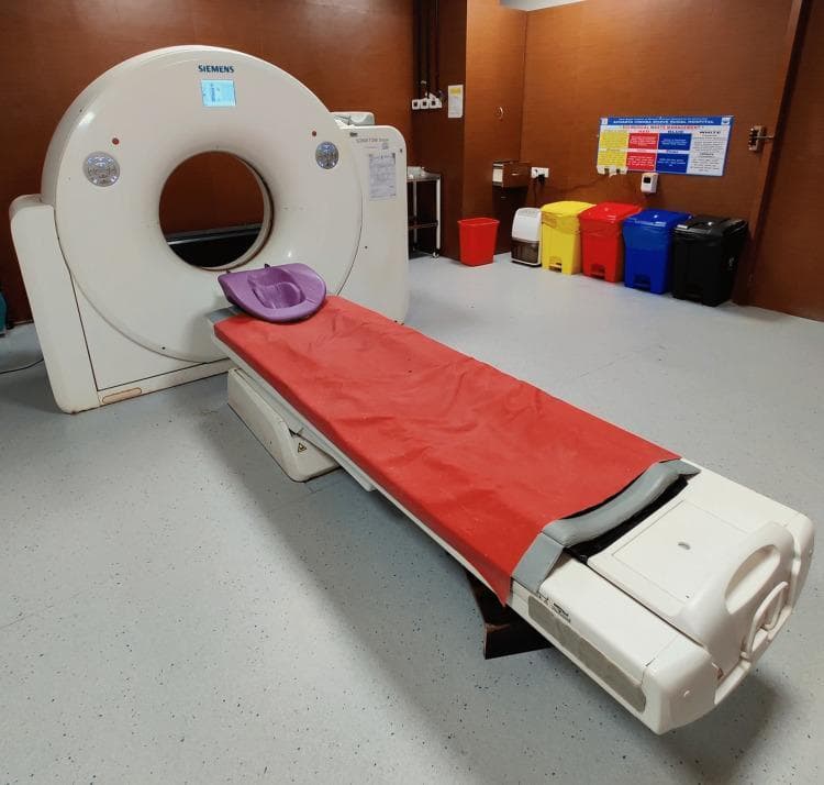 A clinical photograph of a modern Computed Tomography (CT) scanner set in a diagnostic imaging suite. The image features a white Siemens CT gantry with a large central bore and integrated digital display at the top. A motorized patient table extends from the gantry, equipped with a red protective cover and a contoured purple headrest. The clinical environment is further detailed by the presence of a color-coded waste segregation system consisting of red, yellow, blue, and black disposal bins along the back wall, beneath a medical facility poster. This visual provides educational context for diagnostic radiology equipment, the mechanics of cross-sectional imaging (slices), and the standard clinical setup required for trauma, oncology, and neurology diagnostics. The configuration illustrates the essential components used in human medical imaging for identifying internal pathologies such as fractures, tumors, or vascular abnormalities.