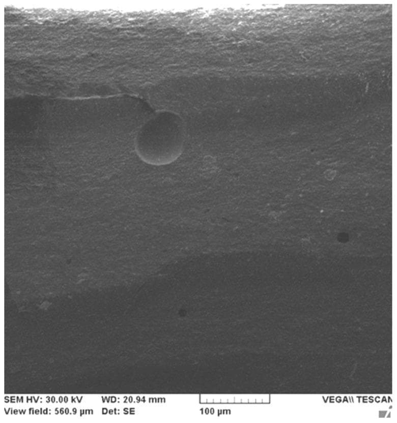 This Scanning Electron Microscopy (SEM) image displays the microstructural topography of a dental restorative composite (Filtek Z250-4). The imaging reveals a heterogeneous surface texture characteristic of a hybrid composite material. A prominent horizontal microcrack, approximately 150 µm in length, is visible in the upper quadrant, where its linear propagation is physically obstructed by a large, spherical micrometer-sized filler particle (approximately 40 µm in diameter). This interaction demonstrates the 'crack-blocking' mechanism which enhances the mechanical properties and fracture toughness of the material. Additional surface irregularities include small dark circular pits, identified as microfiller pull-outs, where filler particles have been dislodged from the resin matrix. The granular background represents the surrounding matrix embedded with varied sizes of filler particles. The image provides clinical relevance regarding the durability and stress-resistance of dental biomaterials under compressive load. A 100 μm scale bar is provided for dimensional reference.