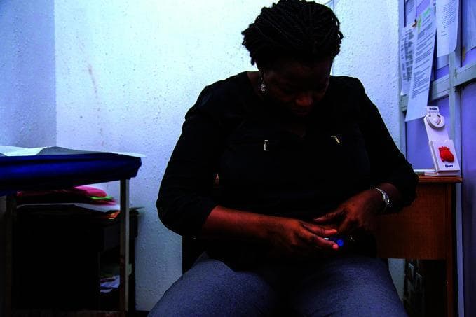 This clinical photograph illustrates a patient self-administering medication, specifically an insulin injection, using an insulin pen device. The image shows a woman in a clinical setting at the Rainbow Specialist Medical Center, focusing on the manual dexterity and technique required for subcutaneous injection in the abdominal region. The patient is holding a blue-capped insulin delivery device with both hands, positioned against the abdomen. The clinical environment is evidenced by the presence of a medical examination table with blue draping, anatomical models (a heart model), and clinical documents in the background. This visual serves as an educational example of diabetes self-management, patient education, and the use of medical devices in a real-world primary care or specialist setting. The photograph emphasizes the practical aspect of chronic disease management and the importance of patient autonomy in endocrinology and metabolic health.