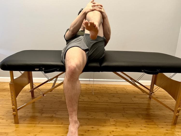 This clinical photograph demonstrates the performance of a Modified Thomas test, a common orthopedic physical examination maneuver used to assess the flexibility of the hip flexors. The patient is positioned supine on a treatment table. One leg (the contralateral limb) is flexed at the hip and knee, with the patient using their hands to pull the knee toward the chest to stabilize the pelvis and flatten the lumbar spine. The test limb is allowed to extend. This visual assessment is used to identify potential muscular tightness or contractures: hip flexion in the test limb suggests psoas involvement, knee extension suggests rectus femoris tightness, and hip abduction or rotation may indicate iliotibial band (ITB) restrictions or tensor fasciae latae (TFL) involvement. This diagnostic maneuver is critical in sports medicine and physical therapy for evaluating hip and knee pathology during a musculoskeletal examination.